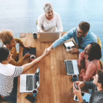 A group of people seated around a table, two of them shake their hands showing Member onboarding
